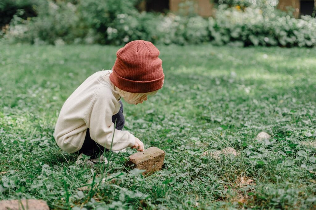 Demo Post 2 Full body side view of playful kid wearing hat squatting on grassy lawn while exploring small stone against blurred background