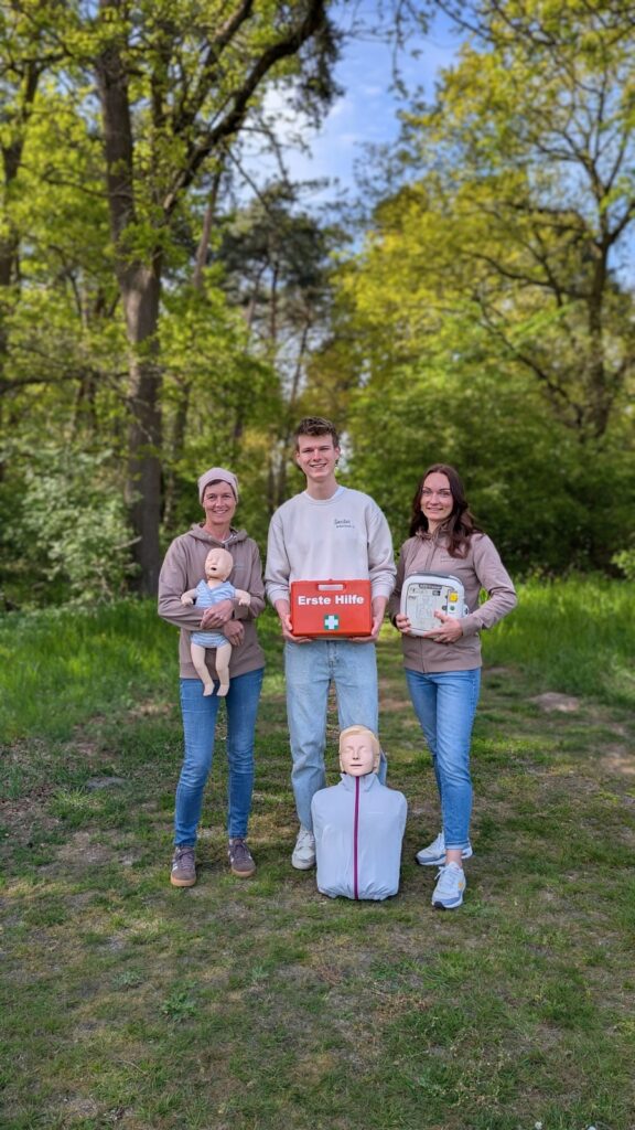 Sanitas - Erste Hilfe Drei Kursleitende mit Erste-Hilfe-Koffer, AED und Übungspuppen auf einer Waldwiese